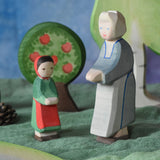 An Ostheimer grandmother figure placed next to a little red riding hood figure in a small world play scene. There is a green coloured decorative cloth covering the surface beneath them and an apple tree and large birch tree in the background.  These wooden figures are part of a wide range of expertly hand crafted wooden toys here at Babipur.