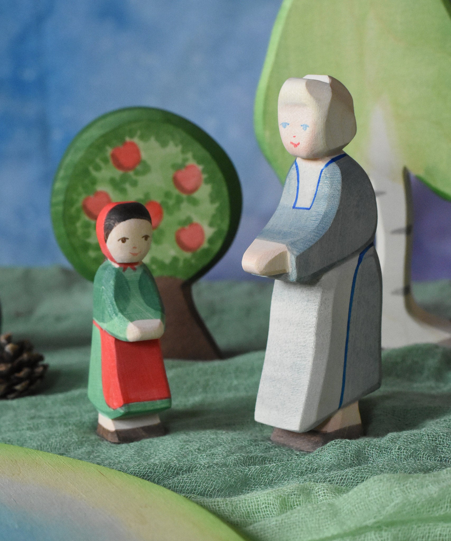 An Ostheimer grandmother figure placed next to a little red riding hood figure in a small world play scene. There is a green coloured decorative cloth covering the surface beneath them and an apple tree and large birch tree in the background.  These wooden figures are part of a wide range of expertly hand crafted wooden toys here at Babipur.