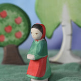 An Ostheimer wooden little red riding hood toy figure placed in the middle of a small world play scene. There is an apple tree and large birch tree in the background with a green coloured decorative scarf underneath them. These wooden figures are part of a wide range of expertly hand crafted wooden toys here at Babipur.