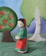 An Ostheimer wooden little red riding hood toy figure placed in the middle of a small world play scene. There is an apple tree and large birch tree in the background with a green coloured decorative scarf underneath them. These wooden figures are part of a wide range of expertly hand crafted wooden toys here at Babipur.