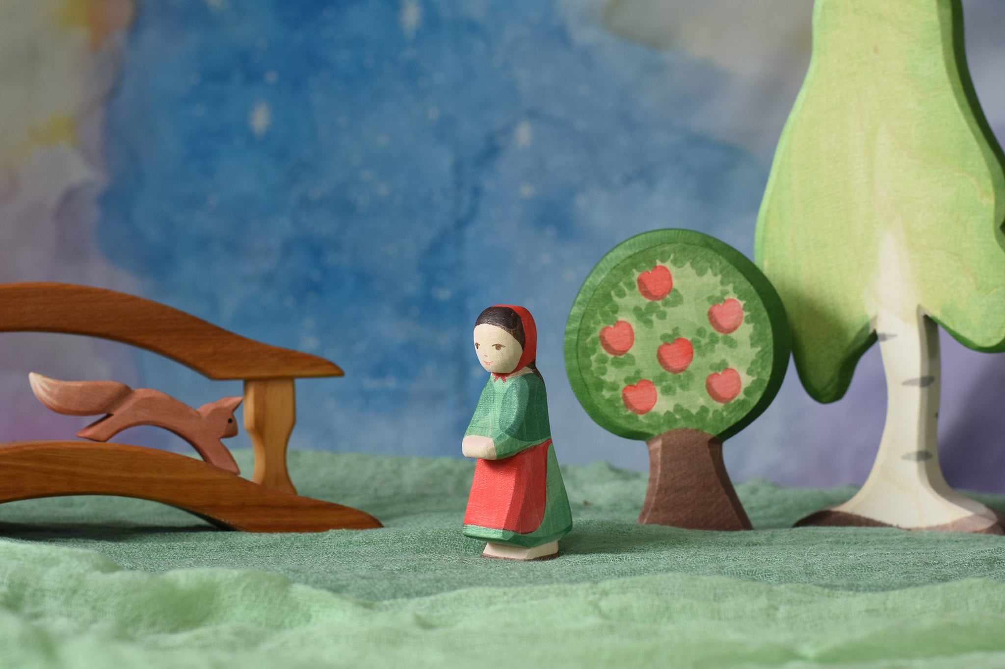 An Ostheimer wooden little red riding hood toy figure placed in the middle of a small world play scene. There is a wooden bridge, squirrel, apple tree and large birch tree in the background with a green coloured decorative scarf underneath them. These wooden figures are part of a wide range of expertly hand crafted wooden toys here at Babipur.