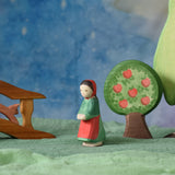 An Ostheimer wooden little red riding hood toy figure placed in the middle of a small world play scene. There is a wooden bridge, squirrel, apple tree and large birch tree in the background with a green coloured decorative scarf underneath them. These wooden figures are part of a wide range of expertly hand crafted wooden toys here at Babipur.