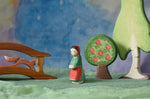 An Ostheimer wooden little red riding hood toy figure placed in the middle of a small world play scene. There is a wooden bridge, squirrel, apple tree and large birch tree in the background with a green coloured decorative scarf underneath them. These wooden figures are part of a wide range of expertly hand crafted wooden toys here at Babipur.