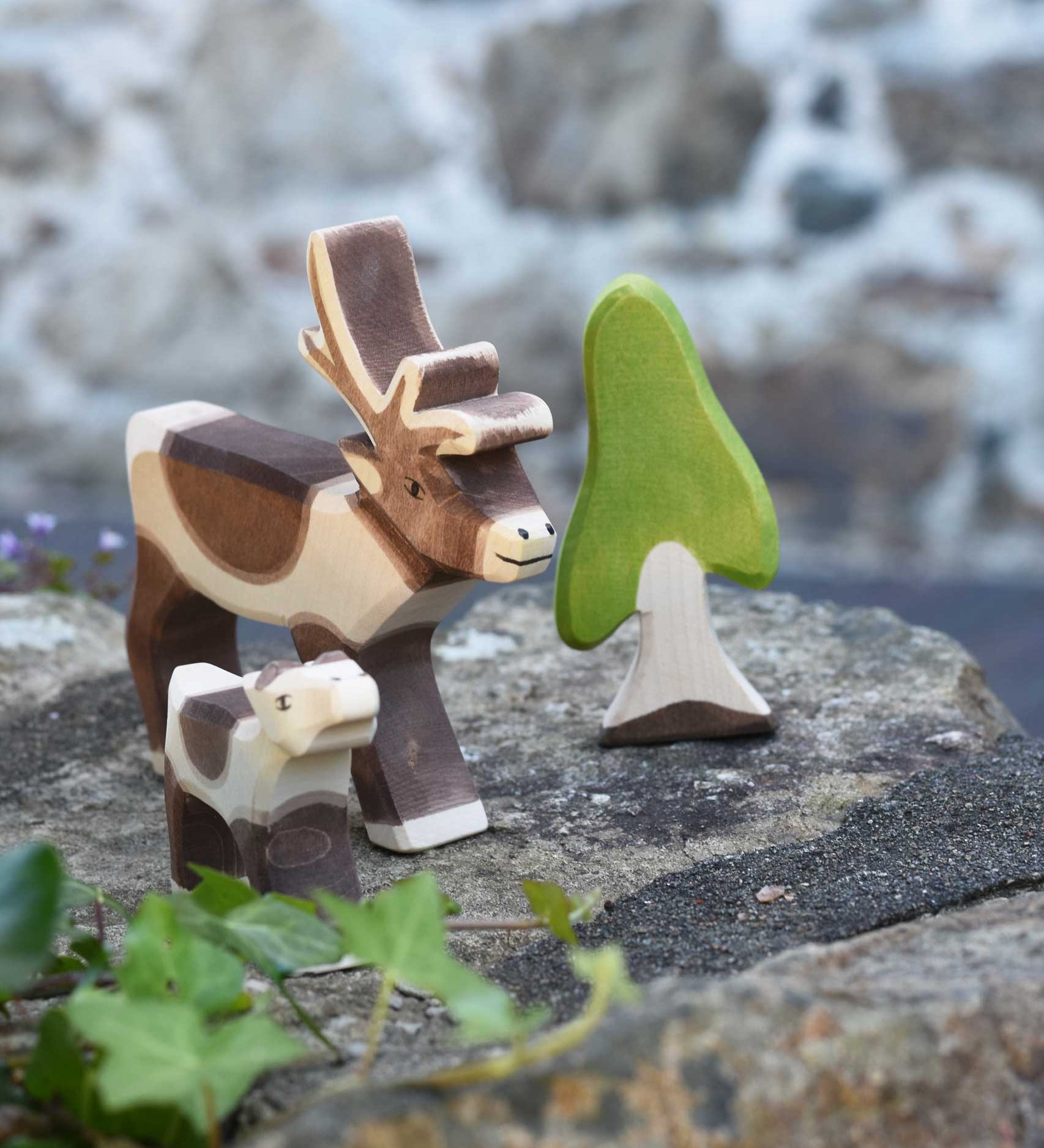 An Ostheimer reindeer and reindeer calf placed on a stone wall outdoors next to an Ostheimer birch tree. Check out our full range of expertly crafted wooden toy figures and scenery here at Babipur.