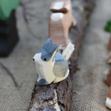 An Ostheimer Wooden Black and White Sitting Rabbit Figure placed on a wooden branch in the middle of a small world play scene. Other Ostheimer toy figures can be seen in the background.