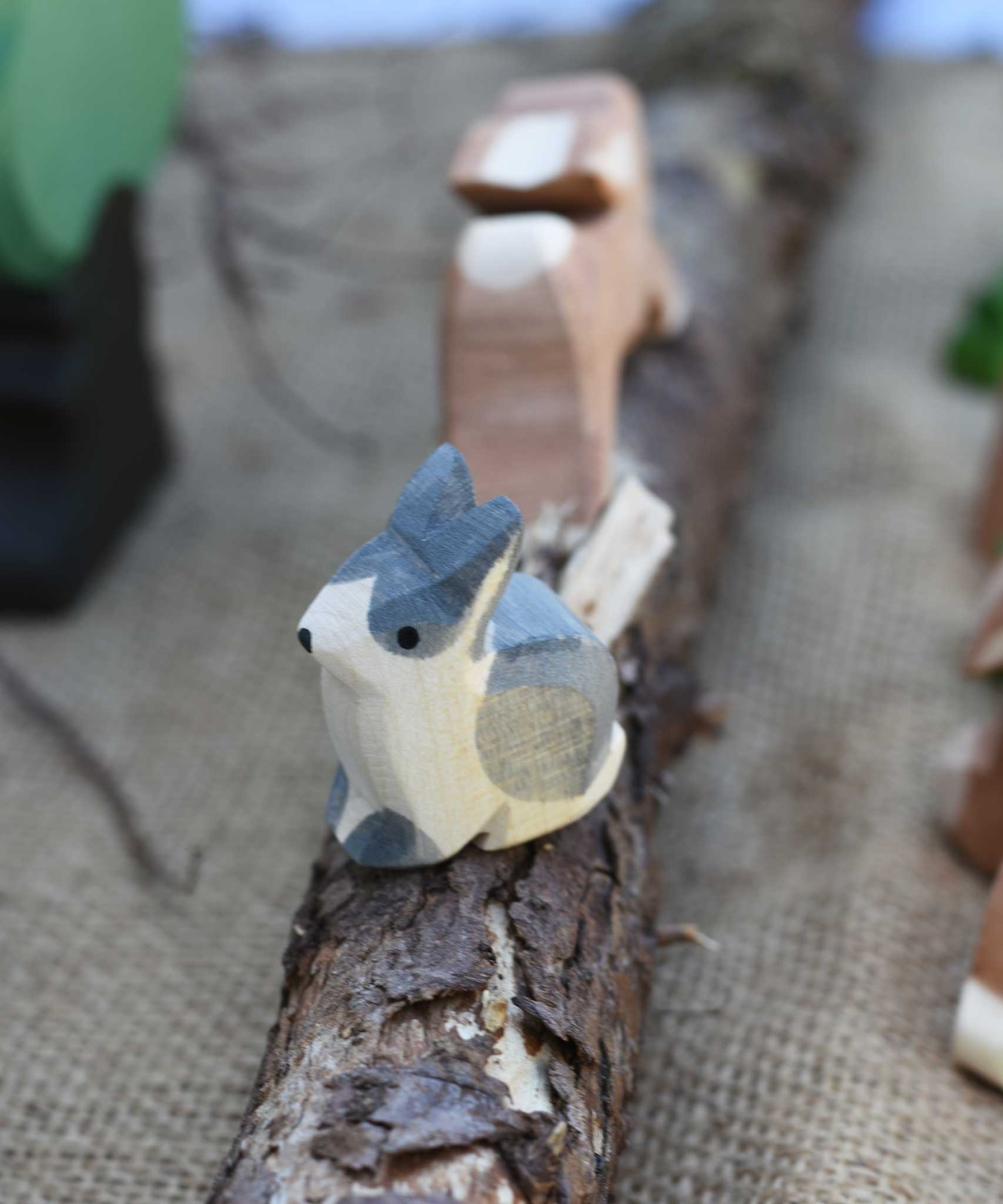 An Ostheimer Wooden Black and White Sitting Rabbit Figure placed on a wooden branch in the middle of a small world play scene. Other Ostheimer toy figures can be seen in the background.