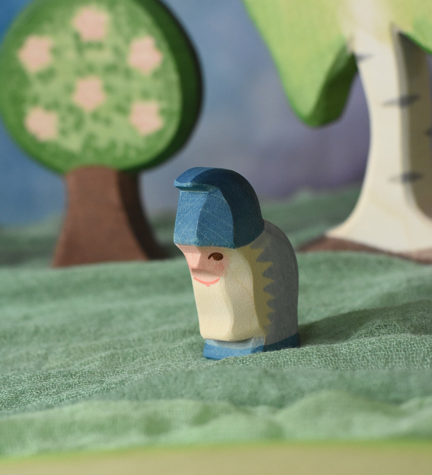 An Ostheimer blue dwarf wooden toy figure placed in a small world play scene. The dwarf figure has a purple coloured outfit with matching hat and a long white beard. The figure has been placed on a green coloured Ostheimer play scarf. This wooden figure is part of a large range of expertly hand crafted, Waldorf inspired, wooden toys here at Babipur.