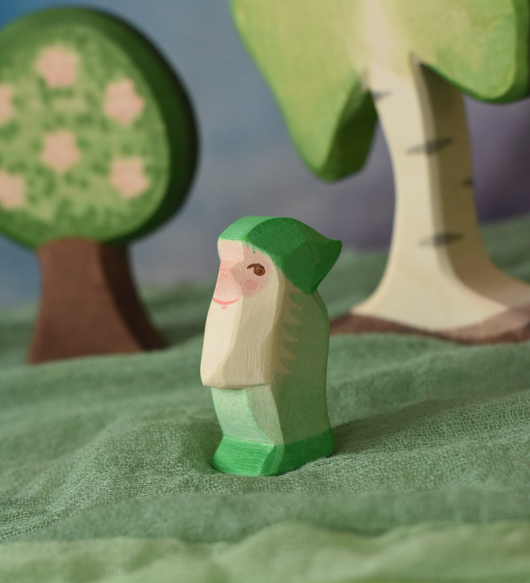 An Ostheimer green dwarf wooden toy figure placed in a small world play scene. The dwarf figure has a green coloured outfit with matching hat and a long white beard. The figure has been placed on a green coloured Ostheimer play scarf. This wooden figure is part of a large range of expertly hand crafted, Waldorf inspired, wooden toys here at Babipur.