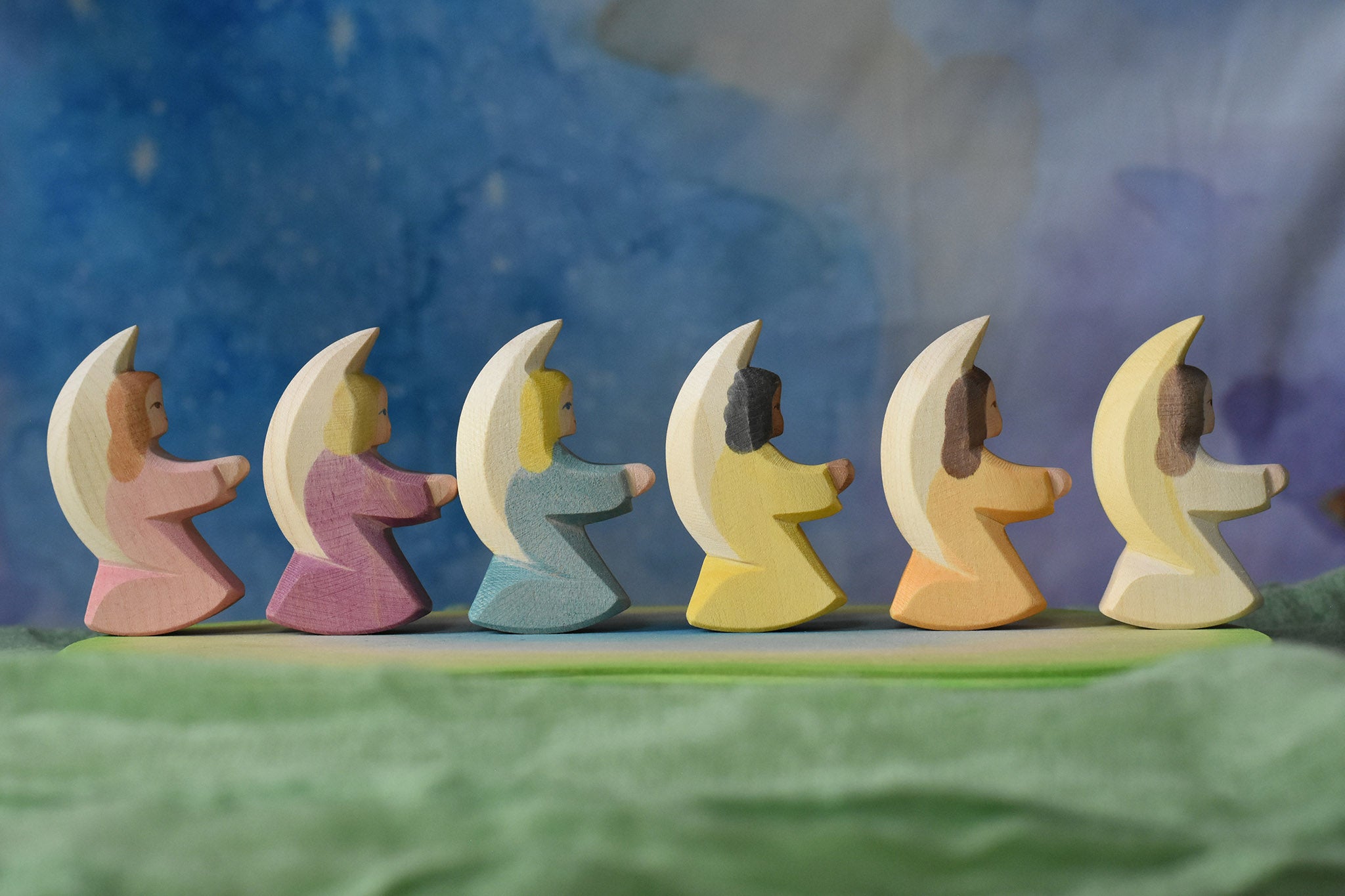 A side profile view of a host of little Ostheimer angels placed in a line on an Ostheimer pond piece. These little angel figures all have different coloured dresses, white, yellow, orange, purple, pink and blue with moon shaped wings.
