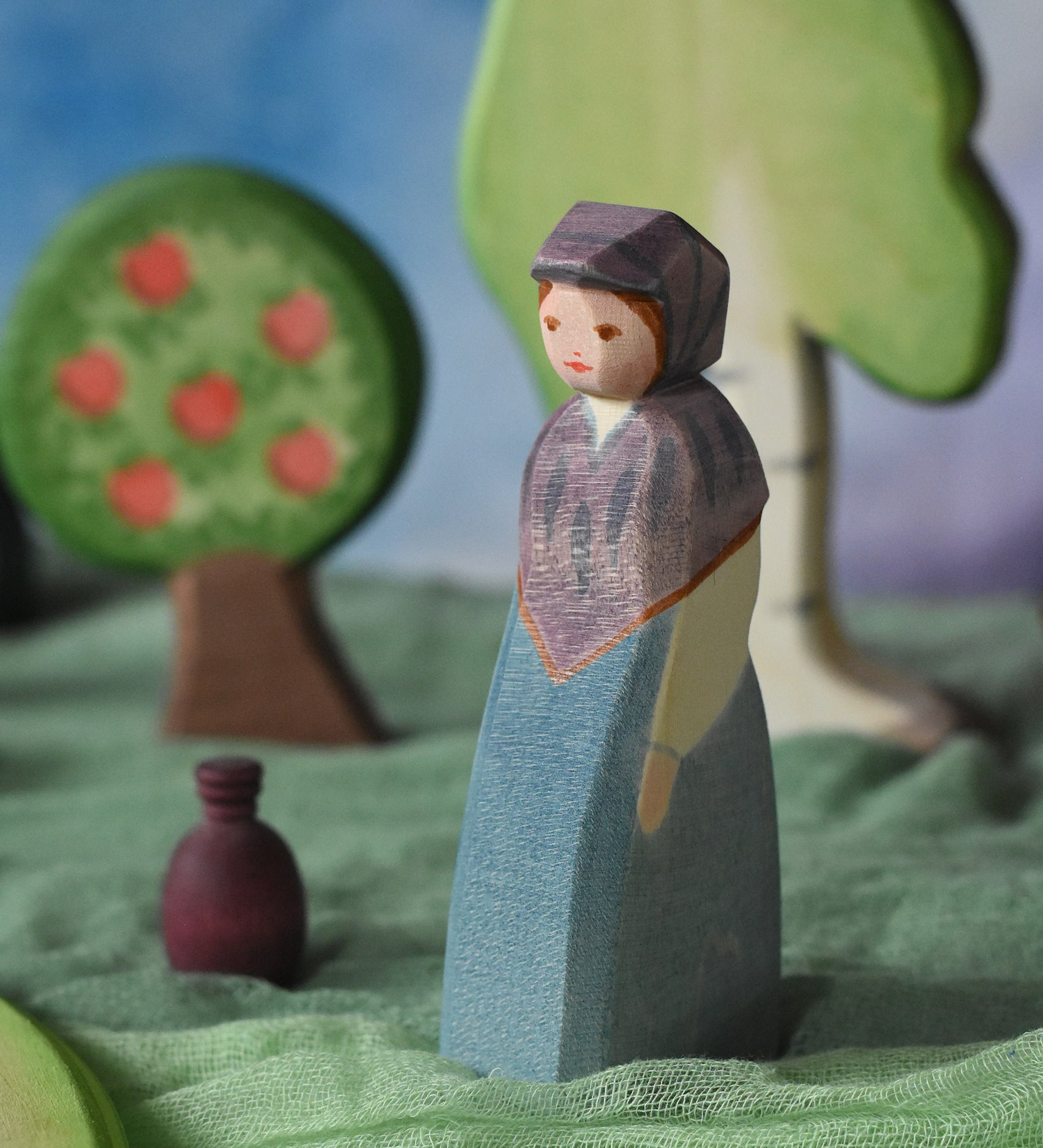 An Ostheimer wooden toy market woman figure placed in a small world scene set up. The figure has a blue coloured dress with a purple shawl and head covering. This wooden figure is part of a large range of expertly hand crafted, Waldorf inspired, wooden toys here at Babipur.
