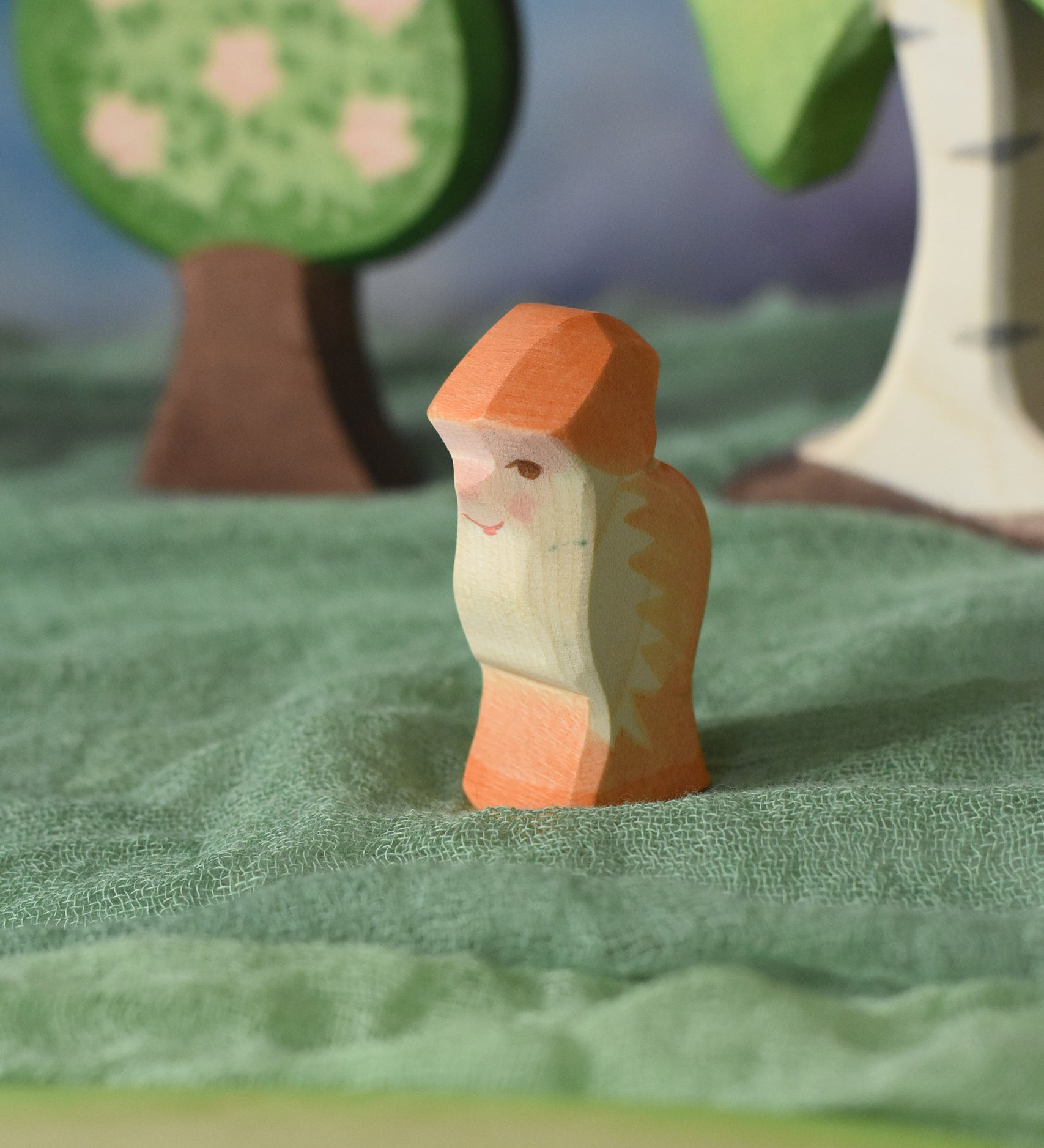 An Ostheimer orange dwarf wooden toy figure placed in a small world play scene. The dwarf figure has an orange coloured outfit with matching hat and a long white beard. The figure has been placed on a green coloured Ostheimer play scarf. This wooden figure is part of a large range of expertly hand crafted, Waldorf inspired, wooden toys here at Babipur.