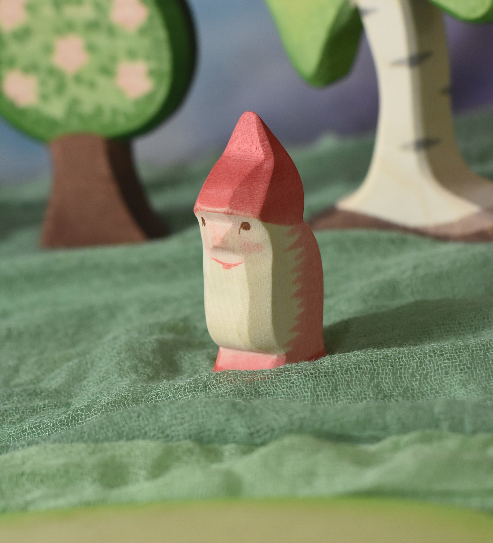 An Ostheimer red dwarf wooden toy figure placed in a small world play scene. The dwarf figure has a red coloured outfit with matching hat and a long white beard. The figure has been placed on a green coloured Ostheimer play scarf. This wooden figure is part of a large range of expertly hand crafted, Waldorf inspired, wooden toys here at Babipur.