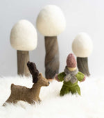 Papoose handmade felt reindeer figure and doll toy stood on a white fluffy blanket in front of 3 white Papoose trees