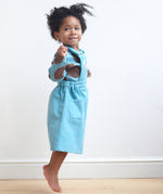 A side image of a girl wearing the Pigeon Organics pinafore dress in turquoise corduroy showing the side pocket detail