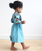 A girl wearing the Pigeon Organics pinafore dress in turquoise corduroy showing the side pocket detail. This dress is available at Babipur.