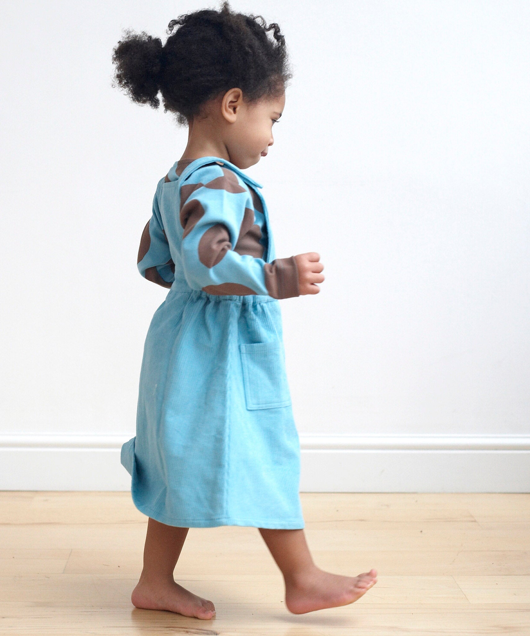 A girl wearing the Pigeon Organics pinafore dress in turquoise corduroy showing the side pocket detail. This dress is available at Babipur.