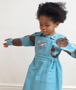 A girl wearing the Pigeon Organics pinafore dress in turquoise corduroy showing the cycling fox embroidery on the front and button fastener detail on the shoulder straps. This dress is available at Babipur.