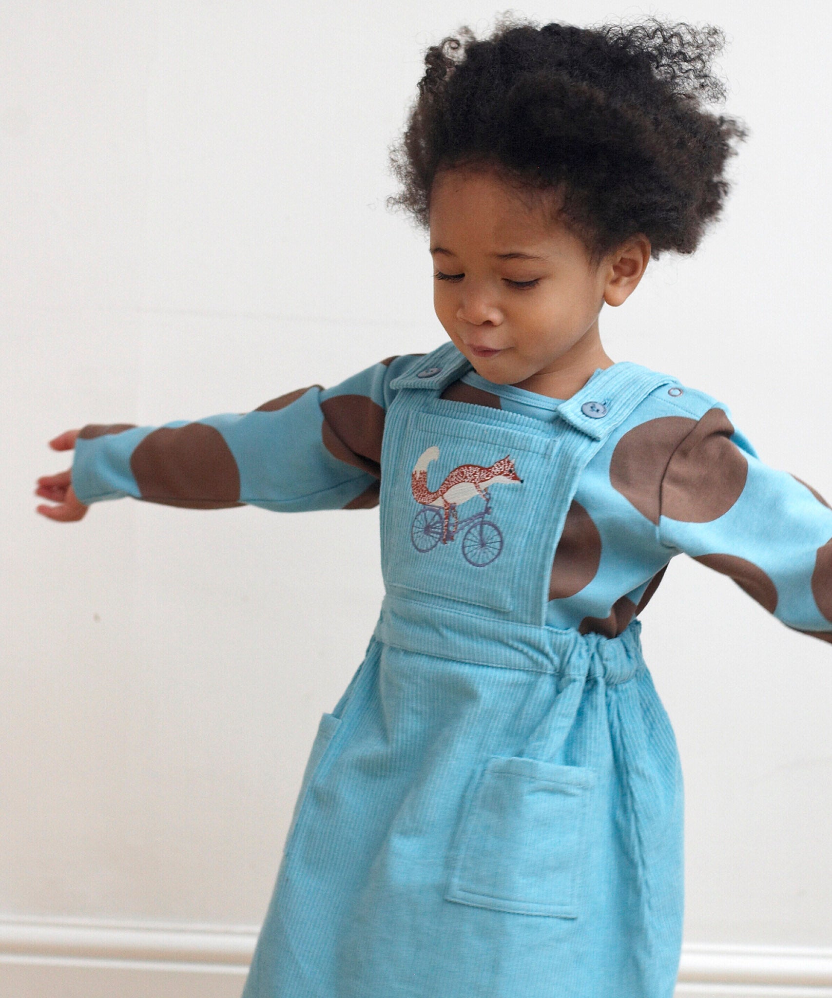 A girl wearing the Pigeon Organics pinafore dress in turquoise corduroy showing the cycling fox embroidery on the front and button fastener detail on the shoulder straps. This dress is available at Babipur.