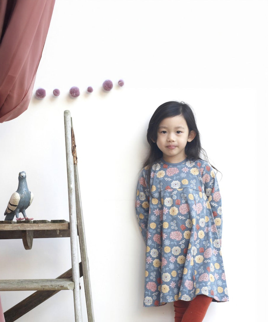 A girl standing up against a wall and wearing the Pigeon Organics pinafore dress - GOTS organic cotton navy long sleeved dress with  multiple prints of multicolour artistic flowers and leaves. Dress available at Babipur.