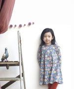 A girl standing up against a wall and wearing the Pigeon Organics pinafore dress - GOTS organic cotton navy long sleeved dress with  multiple prints of multicolour artistic flowers and leaves. Dress available at Babipur.