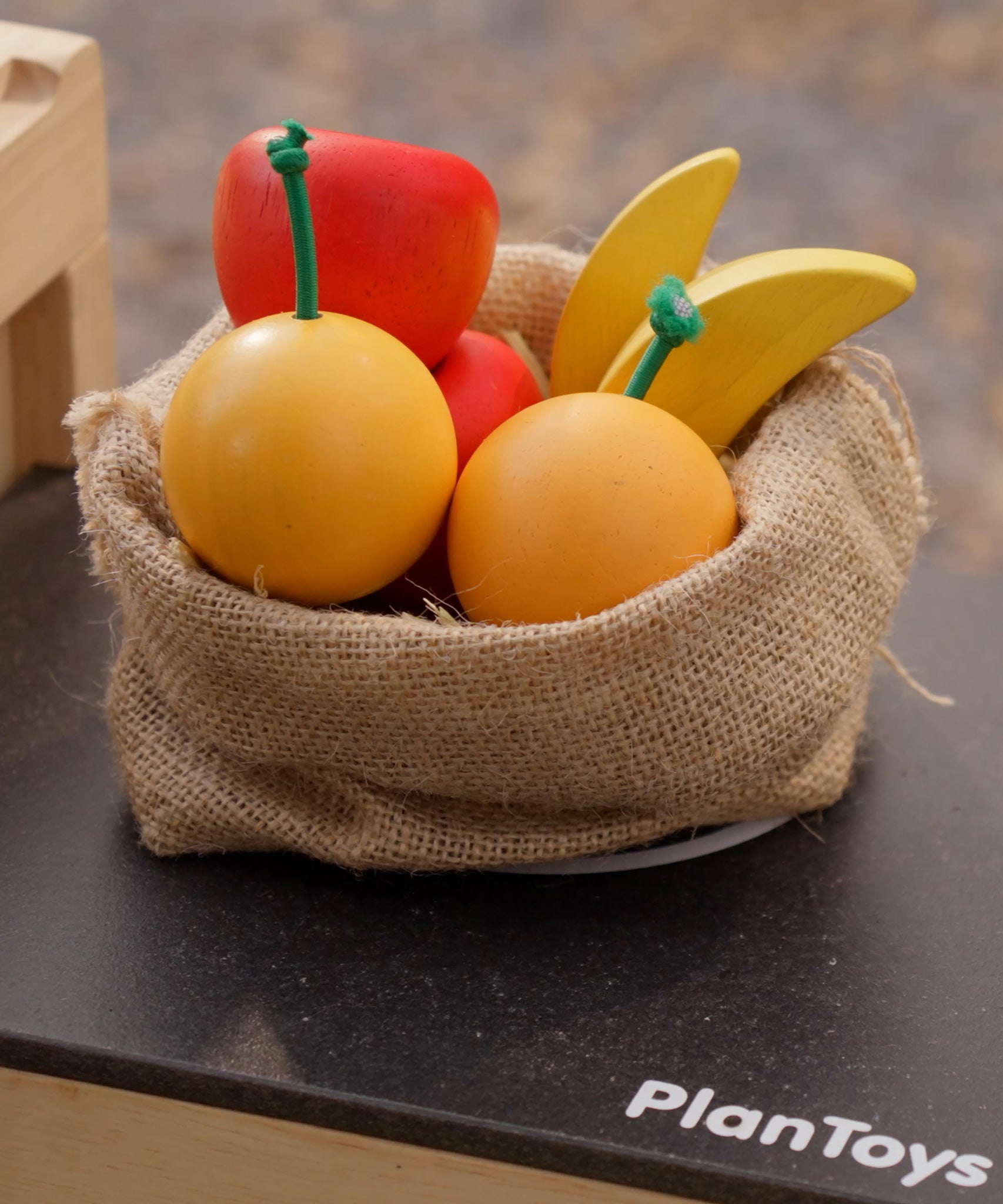 Plan Toys Fruit Produce Set - wooden toy Apples, Oranges & Bananas shown in a jute sack in a play kitchen.