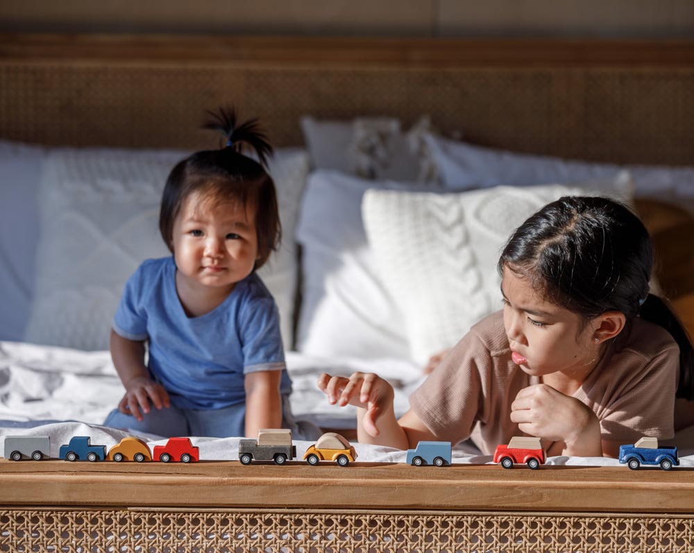 Two children playing with PlanToys vehicles whilst laying on their fronts on a bed.