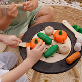 A close up of children playing with the Plan Toys Vegetable Produce Set containing wooden Cucumbers, Onions & Pumpkin play food on a wooden toy grill.