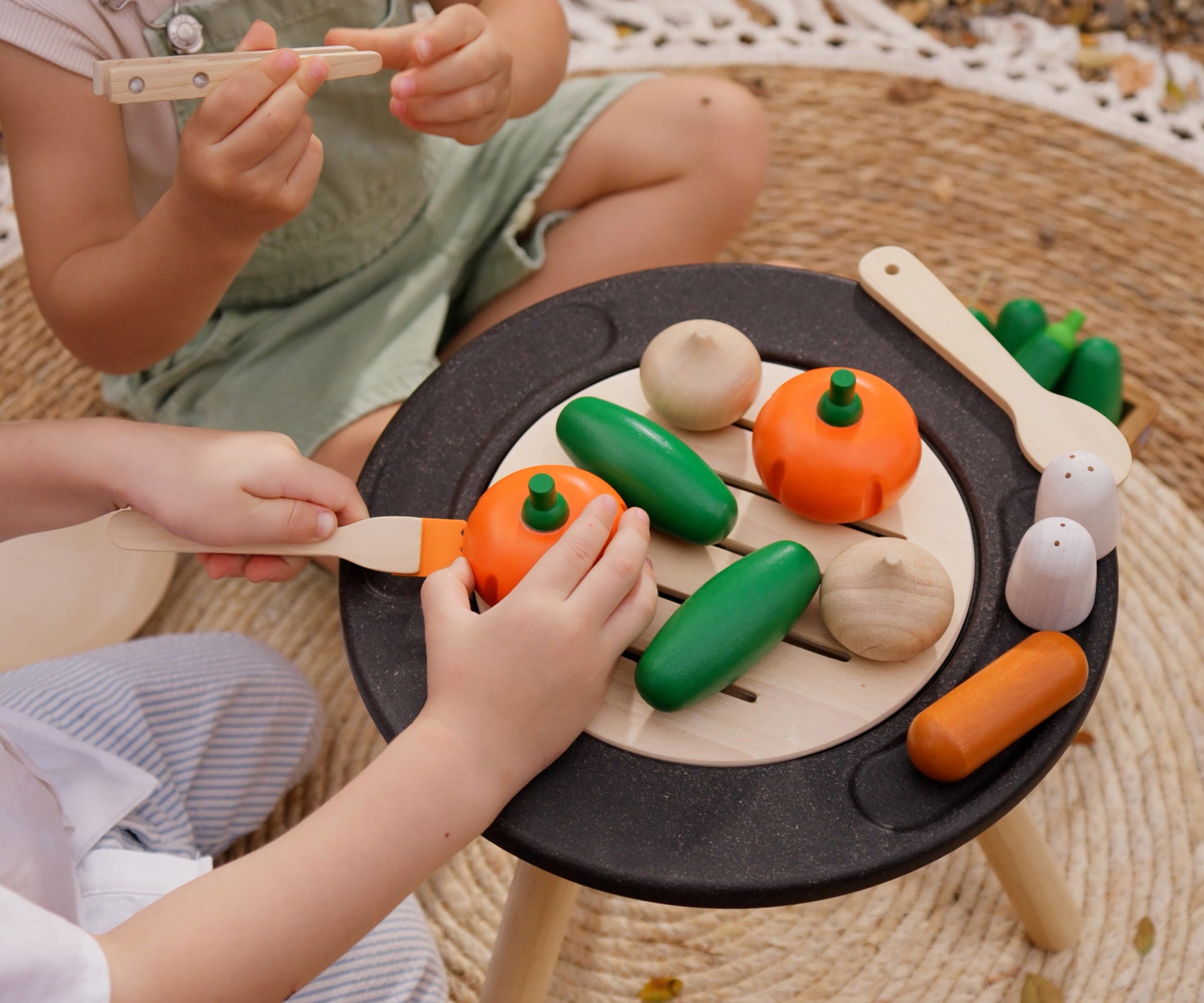 A close up of children playing with the Plan Toys Vegetable Produce Set containing wooden Cucumbers, Onions & Pumpkin play food on a wooden toy grill.