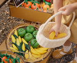 A child placing a wooden toy banana into a basket from the Plan Toys Fruit Produce Set - Apples, Oranges & Bananas.