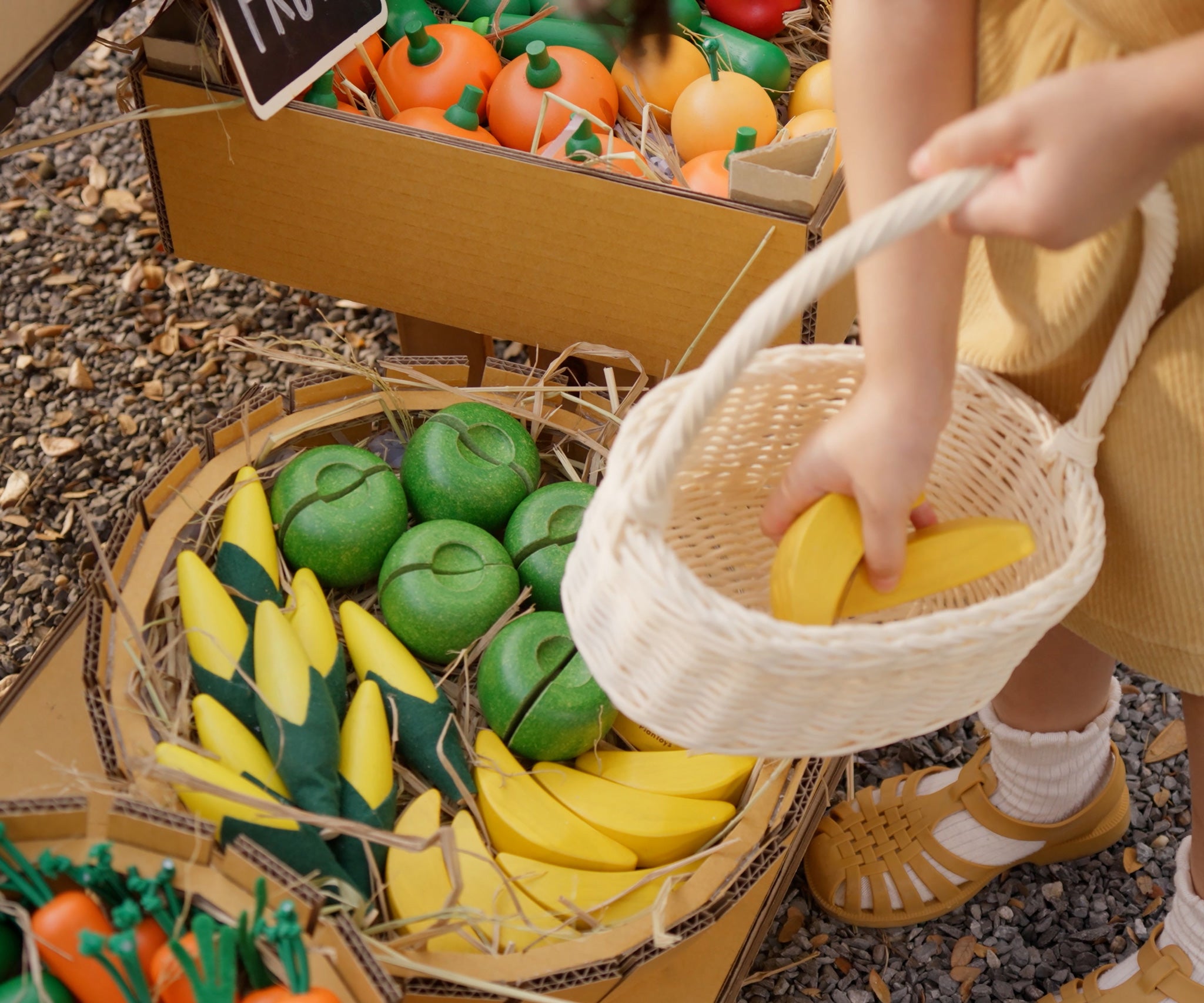 A child placing a wooden toy banana into a basket from the Plan Toys Fruit Produce Set - Apples, Oranges & Bananas.