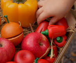 A child's hand reaching into a box that contains the Plan Toys Produce Set - a set of vegetable wooden play food including red peppers, corn and mushrooms.