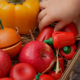 A child's hand reaching into a box that contains the Plan Toys Produce Set - a set of vegetable wooden play food including red peppers, corn and mushrooms.