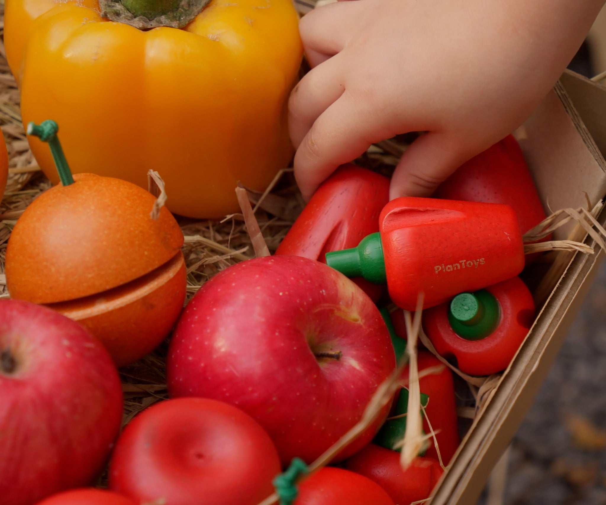A child's hand reaching into a box that contains the Plan Toys Produce Set - a set of vegetable wooden play food including red peppers, corn and mushrooms.