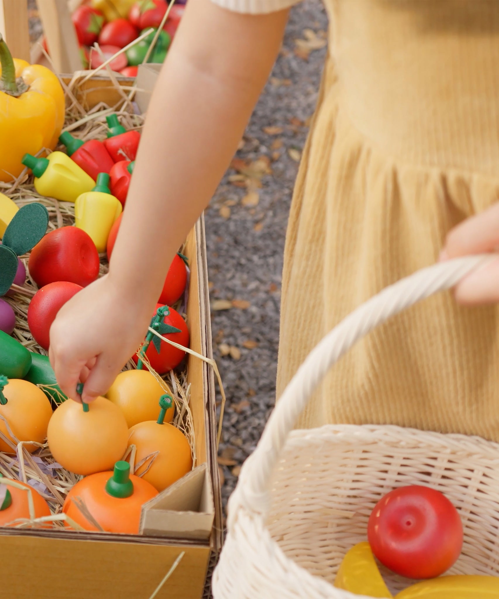 Child reaching out for a wooden toy orange from the Plan Toys Fruit Produce Set. She's holding a basket in one hand that's filled with play food - Apples, Oranges & Bananas