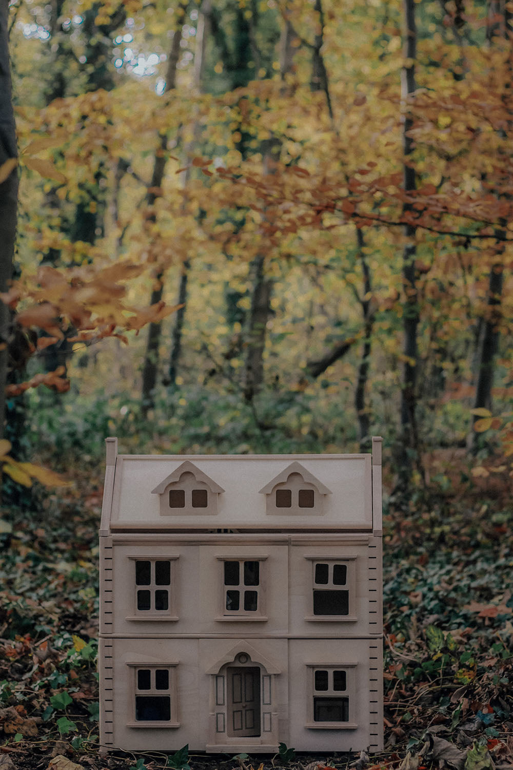A PlanToys Victorian Dolls house in a forest. The ground is covered in fallen leaves.