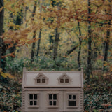 A PlanToys Victorian Dolls house in a forest. The ground is covered in fallen leaves.