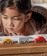 A close up of three vehicles from the PlanToys Mini Cars Set 
laced on a wooden surface. There is a yellow, grey and red car. A child can be seen behind the cars.