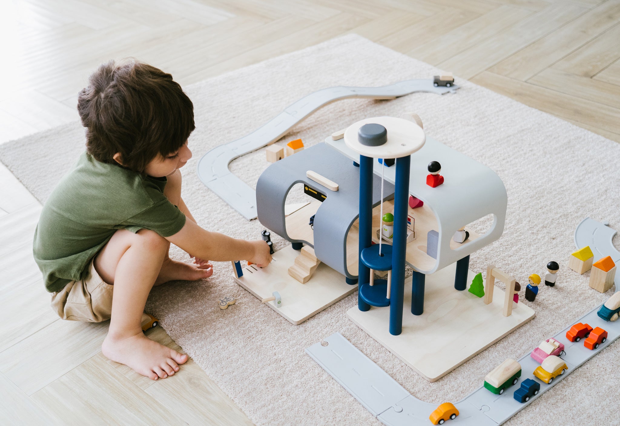 A child crouching down playing with the PlanToys PlanWorld Central Station Wooden toy on a rug.