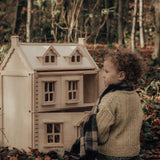 A child sitting on a leaf covered floor in the forest playing with a PlanToys Victorian Dolls House.
