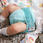 A baby on a bed wearing The Close Pop-In reusable cloth nappy with adjustable velcro, in sky blue.