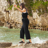 A woman walking on rocks on the beach wearing the black Rapanui jumpsuit. A organic cotton outfit showing the shoulder straps and a matching black belt tie