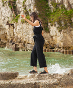 A woman walking on rocks on the beach wearing the black Rapanui jumpsuit. A organic cotton outfit showing the shoulder straps and a matching black belt tie