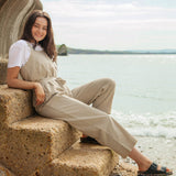 A woman sitting on stone steps on the beach wearing the driftwood Rapanui jumpsuit. A organic cotton outfit in a dark cream colour with shoulder straps and a matching belt tie
