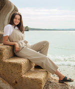 A woman sitting on stone steps on the beach wearing the driftwood Rapanui jumpsuit. A organic cotton outfit in a dark cream colour with shoulder straps and a matching belt tie
