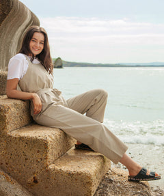 A woman sitting on stone steps on the beach wearing the driftwood Rapanui jumpsuit. A organic cotton outfit in a dark cream colour with shoulder straps and a matching belt tie