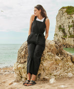 A woman sitting on a boulder on the beach wearing the black Rapanui jumpsuit. A organic cotton outfit with shoulder straps and a matching black belt tie