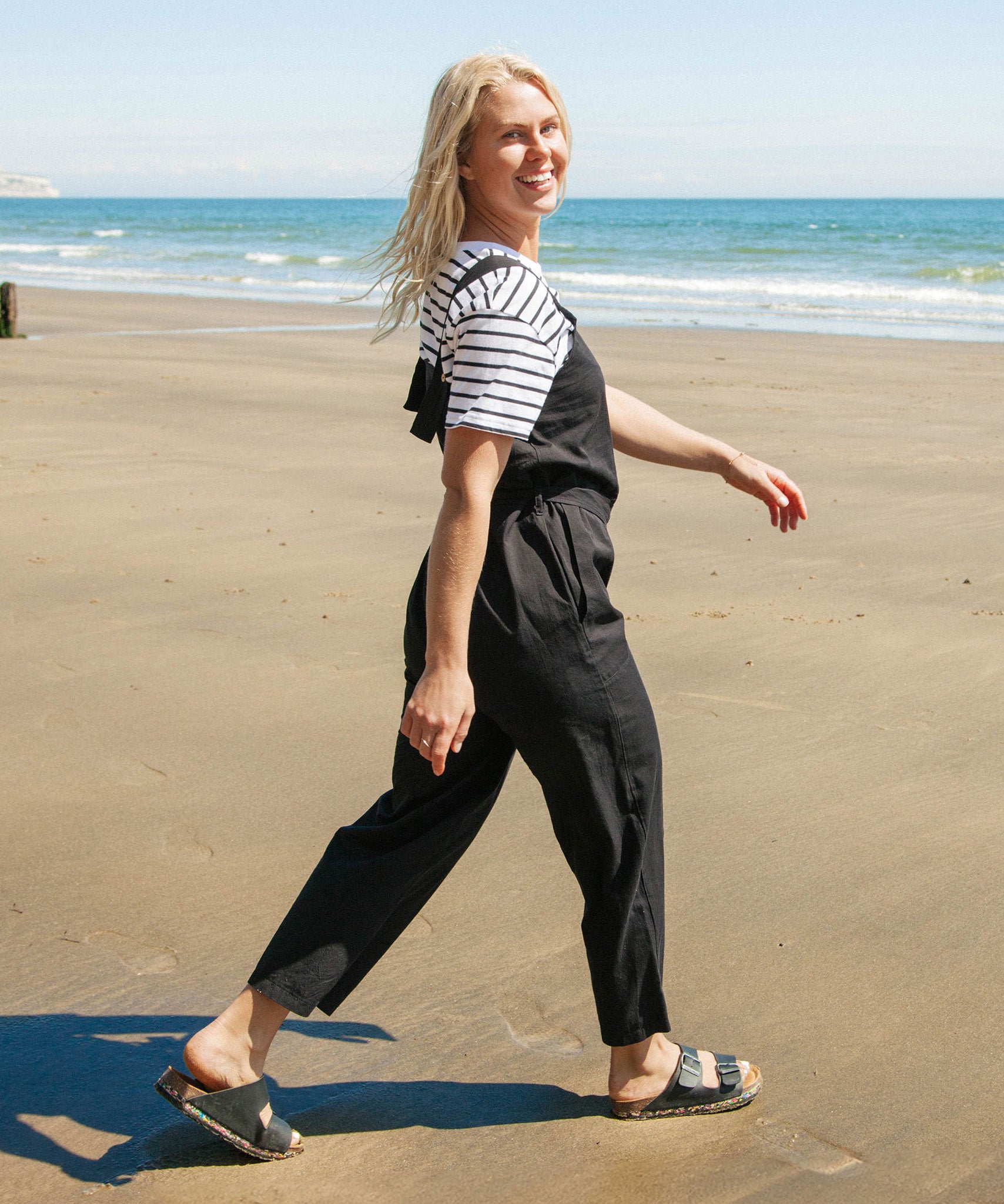 A woman walking on the beach wearing the black Rapanui organic cotton jumpsuit and striped top.