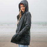 A women standing on the beach wearing the Rapanui maritime coat in black. A long length coat from organic cotton with a PVC -free water resistant coating