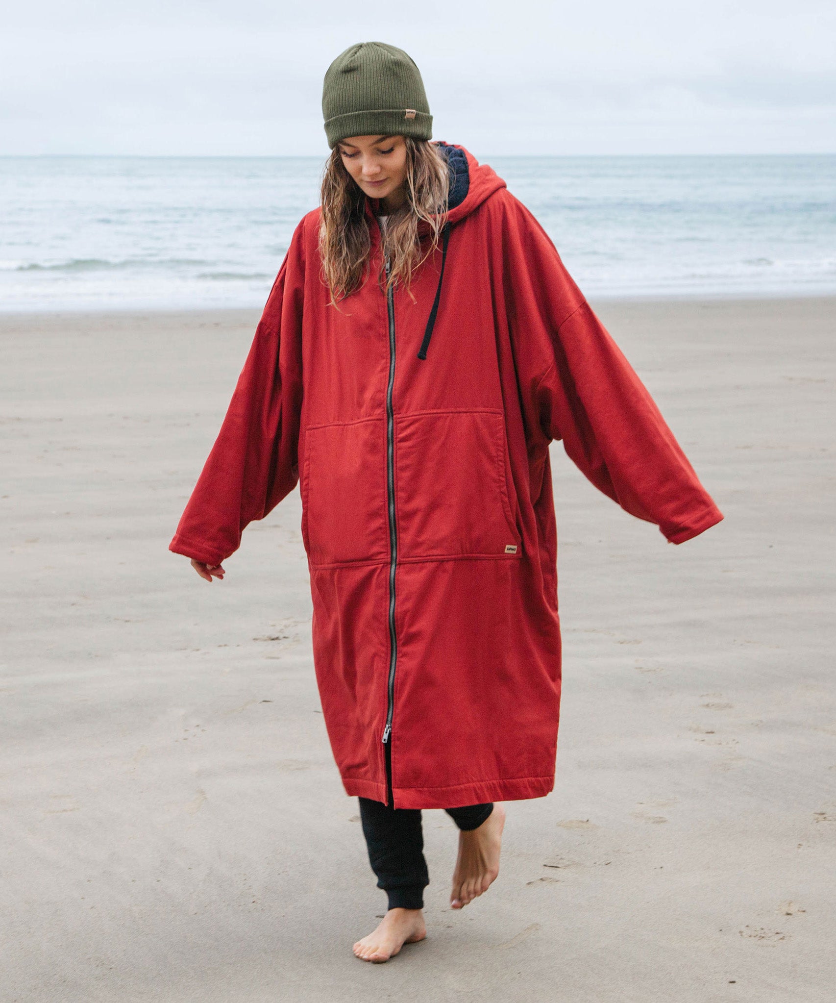 A woman on the beach wearing the Rapanui changing robe in rust colour. A  organic cotton long length changing robe with PFC-free water-resistant coating