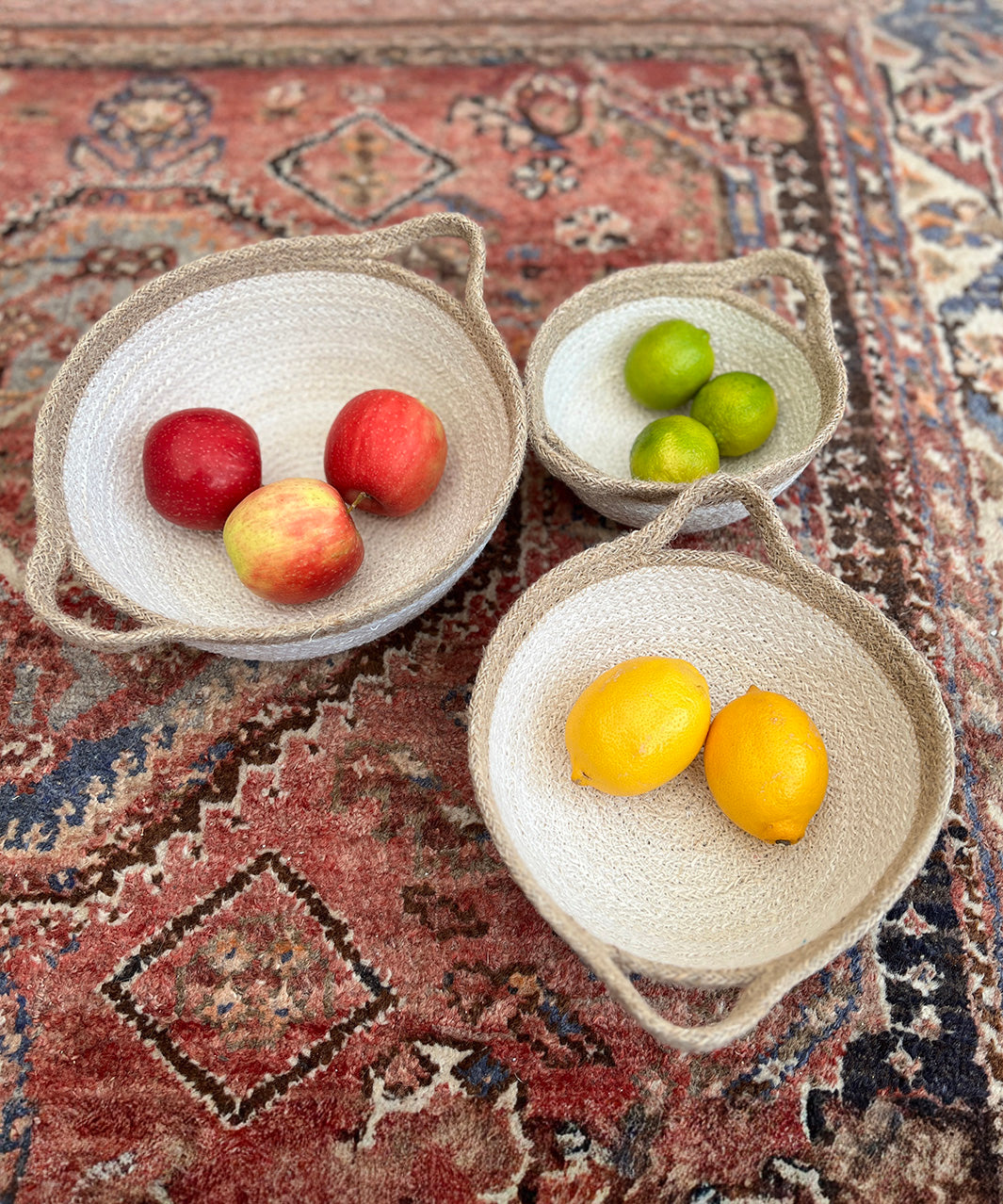 A set of 3 Respiin jute handled bowls on top of a decorated rug with fruit in each bowl
